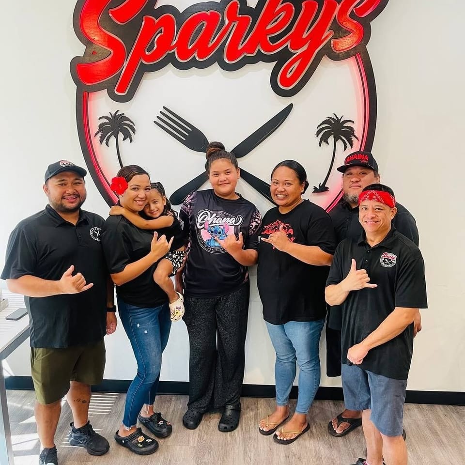 Seven people pose inside Sparky’s restaurant beneath a large illuminated logo featuring crossed fork and knife.