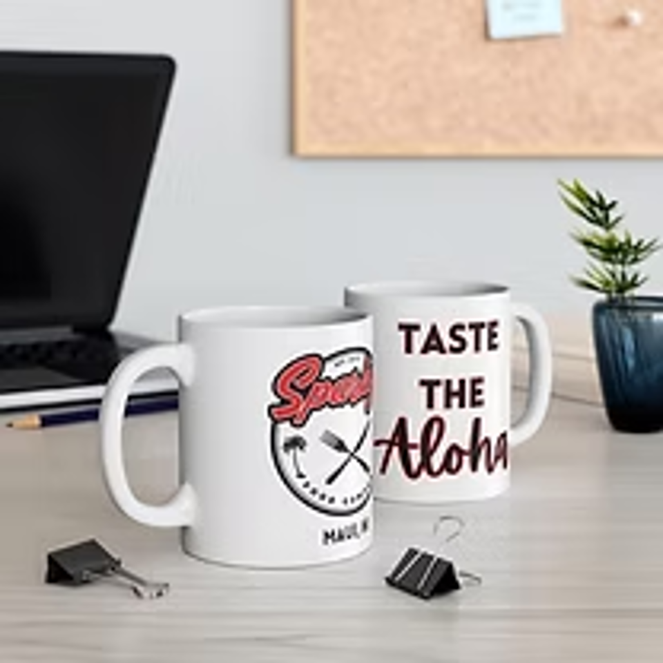 Desk scene featuring a “Taste the Aloha” mug, branded coffee cup, laptop, potted plant, and scattered charging cables.