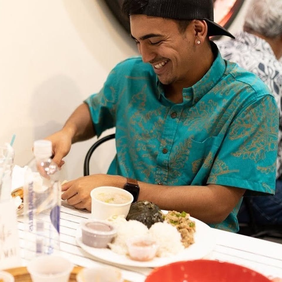 Smiling diner in a turquoise shirt enjoys a plate of rice, meat, and side dishes at a casual restaurant table.